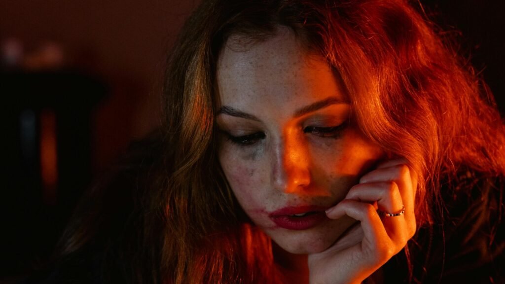 A woman with red hair lies indoors, expression thoughtful, illuminated by smartphone light.